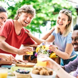 Happy young men and women toasting healthy orange fruit juice at farm house picnic - Life style concept with alternative friends having fun together on afternoon relax time