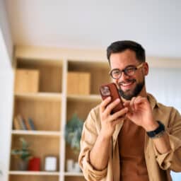 Happy young adult man enjoying time at home, using a mobile phone to browse the internet, read messages, and watch videos, smiling while focused on the screen