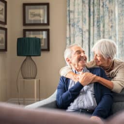 Happy senior woman embracing her husband at home while laughing together. Smiling wife hugging old man sitting on couch from behind. Joyful retired couple having fun at home while looking at each other with copy space.